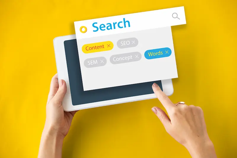 Manos usando una tablet con interfaz de búsqueda que muestra etiquetas como SEO, contenido y palabras clave sobre fondo amarillo